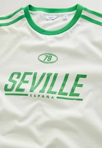 Bílá bavlněná tričko s zeleným kulatým výstřihem, bočními pruhy a opotřebovaným potiskem "SEVILLE 79" na přední straně. Měkká textura, ležérní design.