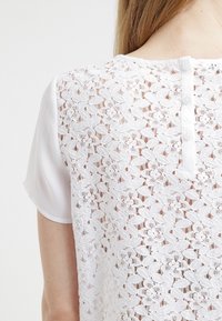 Blusa bianca con retro in pizzo a motivo floreale, maniche corte in satin e chiusura a bottoni al collo. Design delicato e testurizzato.