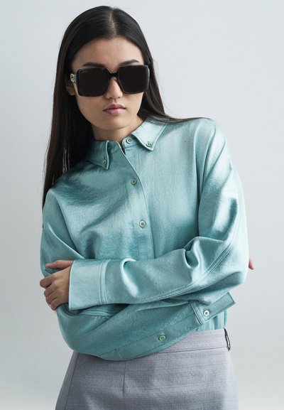 Μια ανοιχτή τυρκουάζ σατέν πουκαμίσα με μακριά μανίκια, υπερμεγέθη μανσέτα και πράσινα κουμπιά, συνδυασμένη με γκρι υφασμάτινο παντελόνι και υπερμεγέθη σκούρα γυαλιά ηλίου.