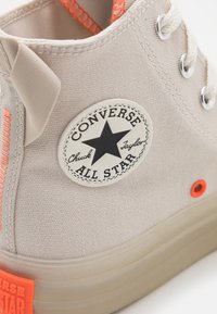 Beige canvas högsko med en orange ögla och logopatch, med en texturerad yta och en genomskinlig sula.