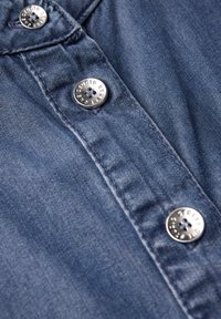 Denim-paita, jossa on sinertävä sävy, metalliset painonapit kaiverretulla tekstillä sekä näkyvät ompelusarjat kankaan pinnalla.