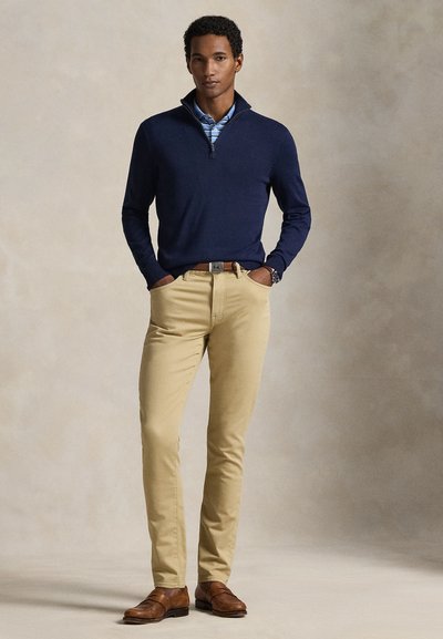 Σκούτερ μπλε ναυτικού τύπου με φερμουάρ στον λαιμό, ανοιχτό μπλε ριγέ πουκάμισο από κάτω, μπεζ chinos, καφέ δερμάτινα παπούτσια και καφέ ζώνη. Απλός και εφαρμοστός σχεδιασμός.