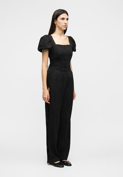 Γυναίκα που φοράει μαύρο σύνολο με κοντά φουσκωτά μανίκια, τοπ με τετράγωνο ντεκολτέ, ψηλόμεσο παντελόνι με φαρδιά πόδια και μαύρα επίπεδα παπούτσια σε μονόχρωμο φόντο.