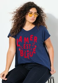 Námořnická modrá krátkosrstá tričko s červeným textem "LA MER EST BLEUE." Měkká tkanina, uvolněný střih, nošeno s tmavými tečkovanými kalhotami a žlutými slunečními brýlemi.