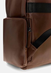 Sac à dos en cuir marron avec une finition texturée, doté d'une sangle noire, d'une poche zippée et d'un détail de couture contrastant.