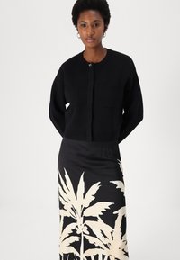 Zwart gebreid vest met zakken, gedragen over een lange rok met grote witte palmbladprint op een zwarte achtergrond.