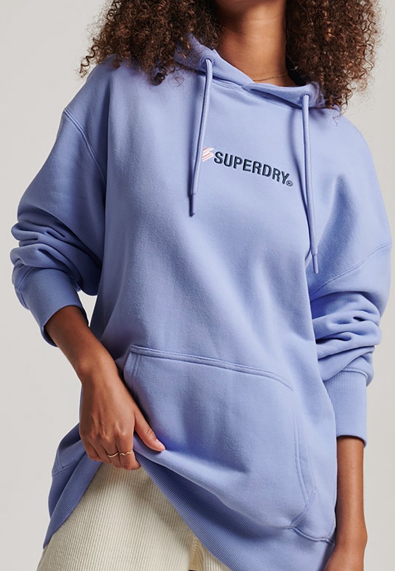 Personne portant un sweat à capuche Superdry violet clair oversize avec poche avant et cordons, associé à un pantalon clair, main reposant sur l'ourlet du sweat.