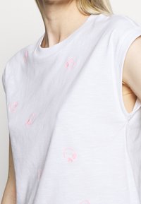 Primer plano de una persona que lleva una camiseta blanca sin mangas, con pequeños bordados de auriculares rosas dispersos por la tela.