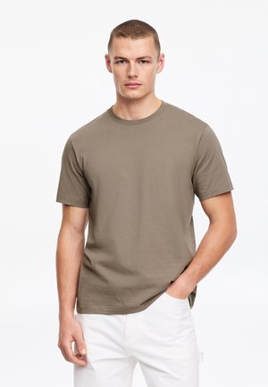 Jeune homme portant un t-shirt à manches courtes vert olive et un pantalon blanc, debout avec une main dans la poche sur un fond uni.