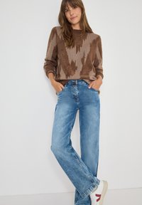 Vrouw in een bruin gebreid patroontrui, blauwe wijde jeans en witte sneakers met rode details, staand tegen een witte muur met haar handen in de zakken.