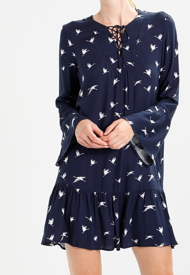 Laivastonsininen mekko, jossa on valkoisia lintukuosisia, V-pääntie nauhakiinnityksellä, kellotetut hihat ja levenevä helma. Valmistettu pehmeästä materiaalista.