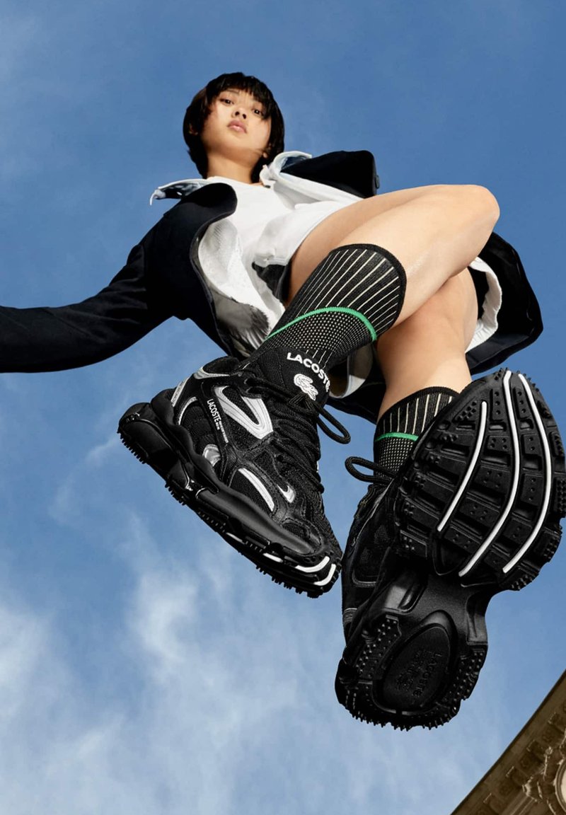 Les chaussures de sport noires Lacoste présentent un dessus texturé, des accents blancs, des lacets proéminents et une semelle robuste. Des chaussettes à rayures vertes complètent le look.