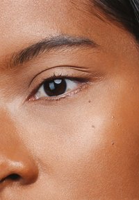Nahaufnahme eines braunen Hauttons mit glatter Textur, dunklen braunen Augen, dezenten Wimpern und einigen kleinen Wassertropfen auf der Haut.