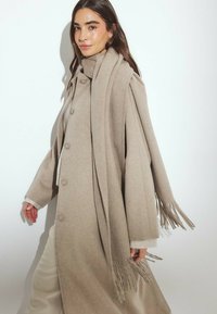 Béžový vlněný kabát s velkými knoflíky, širokými rukávmi a zodpovedajúcim šálom s strapcami. Hladká textura, dĺžka po kolená.