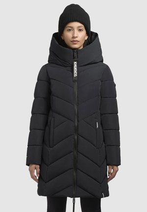 Schwarze Pufferjacke mit hohem Kragen, Zickzack-Steppmuster, Vorderreißverschluss und Seitentaschen. Trägt eine schwarze Strickmütze.