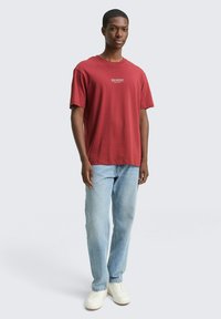 Młody mężczyzna w czerwonej koszulce z grafiką, jasnoniebieskich dżinsach i białych trampkach, stojący na tle jasnoszarego tła.