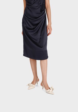 Donna che indossa una gonna midi drappeggiata blu navy e décolleté slingback color crema con grandi fiocchi sulla parte superiore.