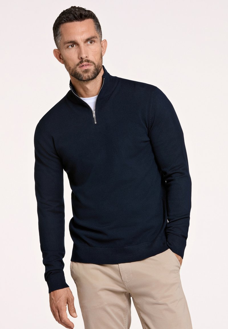 Maglione zip-up blu navy realizzato in un morbido tessuto texturizzato, con maniche lunghe e un design aderente, abbinato a pantaloni beige chiaro.