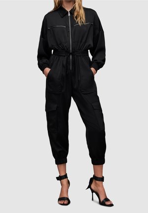 Sort jumpsuit med lynlås foran, to lynlåslommer på brystet, elastisk talje og tapered ankler. Har en afslappet pasform og lange ærmer.