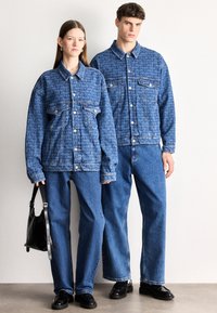 Traper jakna sa uzorkom ponavljajućeg teksta, svijetloplave boje, s kopčama na pritisak, u kombinaciji s odgovarajućim trapericama. Teksturirani materijal i opušteni kroj.