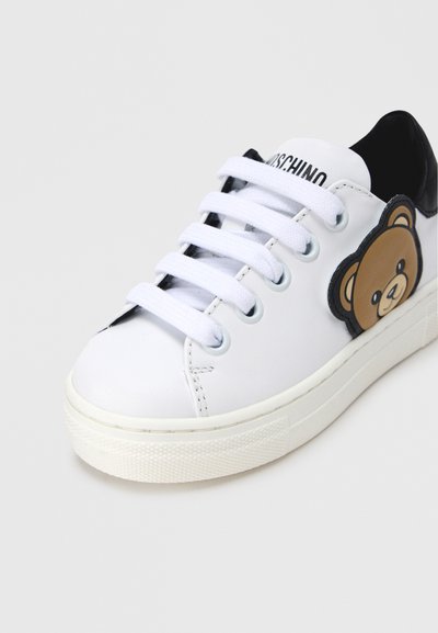 Λευκό δερμάτινο αθλητικό παπούτσι με μαύρες λεπτομέρειες, που έχει ένα καρτούν ταμπελάκι αρκούδας στο πλάι, στρογγυλή μύτη και λαστιχένια σόλα με υφή για καλύτερο κράτημα.