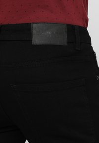 Jeans de mezclilla negra con una etiqueta de cuero negro texturizado. Diseño de cinco bolsillos con costuras en contraste y un corte ajustado. Marca sutilmente visible.