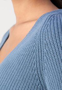 Gros plan sur le cou et l'épaule d'une personne portant un pull tricoté bleu clair avec un motif côtelé texturé.