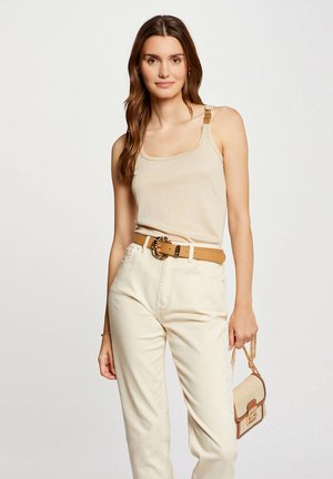 Top de punto beige sin mangas con detalles en tono dorado en los hombros, combinado con pantalones crema de cintura alta y un cinturón marrón claro. Sosteniendo un bolso pequeño.