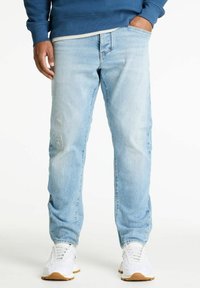 Svetlo modri jeans s sproščenim krojem, ki vključujejo poškodovane detajle in klasičen pet-džepni dizajn. Obuti s belimi športnimi čevlji.