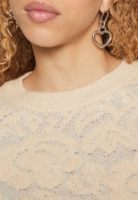 Beiger Strickpullover mit einem strukturierten Muster und silbernen Fäden, kombiniert mit silbernen, herzförmigen Anhängerohrringen.