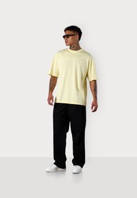 Gelbes Baumwoll-T-Shirt mit kurzen Ärmeln und Rundhalsausschnitt, kombiniert mit schwarzen Hosen und weißen Turnschuhen. Modell trägt dunkle Sonnenbrille.