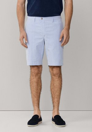 Man draagt lichtblauwe en wit gestreepte korte broek, donkerblauw overhemd en zwarte instapschoenen, staand tegen een effen achtergrond.