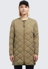 Khaki prešívaná bunda so zapínaním na zips, okrúhlym výstrihom a bočnými vreckami. Štruktúrovaná tkanina má vzor vĺn. Nosená s čiernou čiapkou.