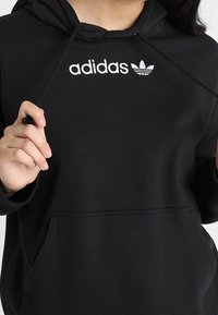Mustapainen huppari, jossa on valkoinen "adidas"-logo ja kolmilehtigrafiikka. Sisältää etutaskun ja nauhat hupussa. Pehmeä kangastunne.