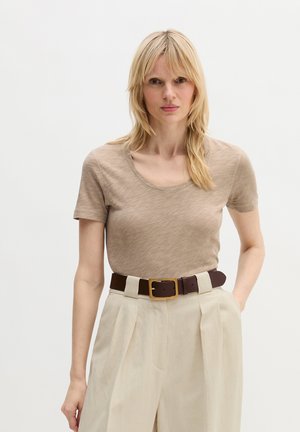 Blondynka stojąca, ubrana w beżową koszulę z krótkim rękawem, kremowe spodnie z wysokim stanem i plisami oraz ciemnobrązowy pasek ze złotą klamrą.