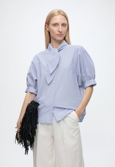 Femme blonde porte un chemisier bleu clair à manches bouffantes avec une cravate et un pantalon blanc à jambes larges, tenant un sac à main noir à franges.