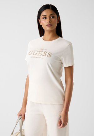 Biały bawełniany T-shirt z beżowym logo „GUESS CALIFORNIA”. Krótkie rękawy, okrągły dekolt i gładka faktura. W połączeniu z białymi spodniami.