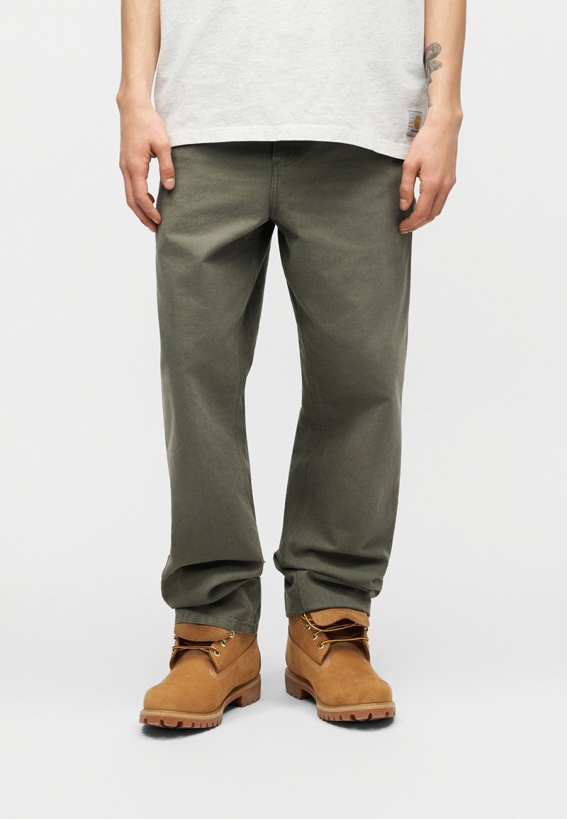 Unterkörper einer Person, die olivgrüne Hosen und sandfarbene Schnürstiefel trägt, stehend vor einem einfarbigen hellen Hintergrund.