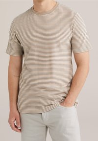 Béžové tričko s krátkým rukávem a tenkými bílými pruhy, s kulatým výstřihem. Měkká textura a přiléhavý střih. Nosené s světle šedými kalhotami.