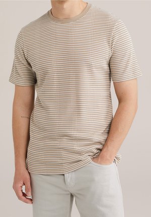 T-shirt beige à manches courtes avec de fines rayures blanches et un col rond. Texture douce avec un design ajusté. Porté avec un pantalon gris clair.