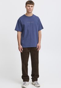 Maglietta di cotone blu navy con scollo a girocollo e maniche corte, con testo ricamato "PLAY ME A MEMORY." Abbinata a pantaloni di velluto a coste marroni.