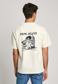 Camiseta de manga corta color crema con una ilustración gráfica en negro de una tienda de discos y el texto "PEPE JEANS" en la parte de atrás.