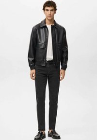 Schwarze Kunstlederjacke mit zwei Fronttaschen, kombiniert mit einem weißen T-Shirt und schwarzen Jeans. Das Modell trägt außerdem schwarze Schuhe.