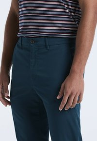 Navyblauwe broek met een gladde textuur, voorzien van een knoopsluiting en zijzakken, gedragen met een gestreept shirt.