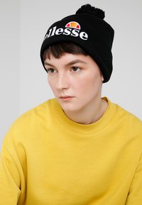 Black knit beanie with a pom-pom on top, featuring white embroidered "ellesse" logo and a circular red and yellow graphic. Worn with a yellow sweatshirt.