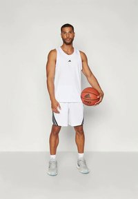 Hvit athletisk tank topp med svart adidas-logo, kombinert med hvite shorts med svarte sidestriper. Holder en basketball. Grå joggesko.