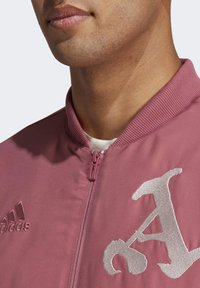 Rosa bomberjacka med hög krage, dragkedja och broderad vit bokstavsdesign. Har Adidas-logotypen på vänster sida. Slät textur.