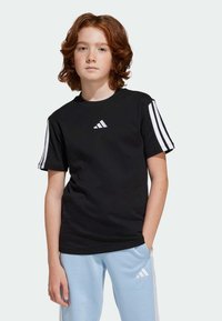 T-shirt en coton noir avec des manches courtes et des rayures blanches, logo blanc en relief sur la poitrine. Porté avec un pantalon de jogging bleu clair.