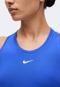Femme portant un débardeur de sport bleu avec un logo Nike blanc, gros plan sur le cou et le bas du visage légèrement tournés vers la gauche.