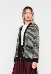 Woman wearing black and white houndstooth cardigan with gold buttons, white top, and burgundy skirt, looking to her right against white background.